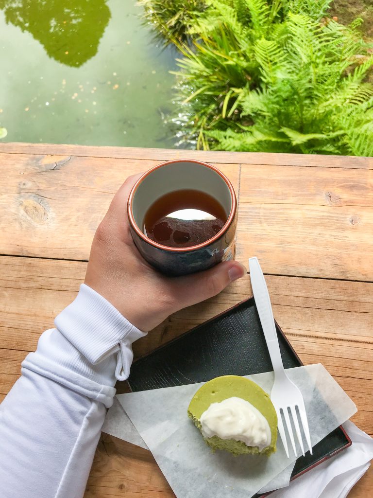 Author holding a cup of Hojicha with a Green Tea Cheese Cake overlooking the pond at the Japanese Tea Garden in San Francisco